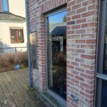 View from inside a building looking out at a small, walled courtyard where tall grass grows freely around a modern, round plant pot, with the visible window and door potentially requiring windows and door repair in Austin